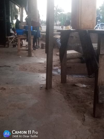 The image captures an outdoor scene, likely a market or informal trading area, in Unguwan Rimi, Nigeria. Several individuals are present, engaging in daily activities. On the left side, a child stands next to a wooden table where several blue plastic basins are placed, suggesting the handling or sale of goods, possibly food items. Another person is partially visible standing behind this table.
Further to the right, obscured by thick wooden support beams, another person is seated on a low structure. Above them, a large, beige, cylindrical container or tank rests on a raised wooden stand. The ground beneath the structures is an uneven mix of light-colored concrete and darker reddish-brown dirt, with various loose objects and construction materials like bricks scattered around.
The area is characterized by sturdy wooden posts and makeshift tables or stalls. In the background, beyond the immediate structures, a road or street can be discerned, lined with green foliage from trees, indicating an open, possibly roadside, location. The lighting is soft and diffused, suggesting either an overcast day or the early morning/late afternoon, as there are no harsh shadows. The emotions of the individuals are not discernible.
In the bottom-left corner of the image, a camera watermark is visible, displaying the text "CAMON 11 Pro" and "24M AI Clear Selfie."