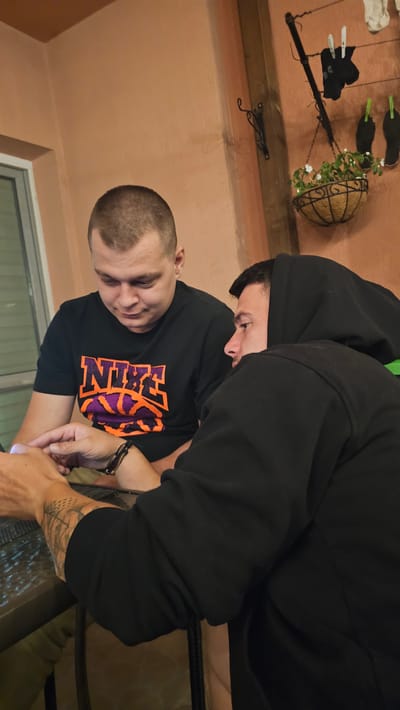 Two adult males are seated at a table, appearing to interact with a laptop. The male on the left, with a shaved head, wears a black t-shirt with an orange and purple Nike logo. The male on the right, wearing a black hooded sweatshirt, is partially obscured, facing left. The background suggests an outdoor or semi-outdoor setting, possibly a patio or balcony, with a peach-colored wall, a window, and a hanging basket with plants. The scene is lit by ambient light. The image appears to be taken in the city of Karlovo, Bulgaria.