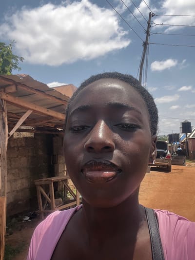A young woman with dark skin and a pink top is taking a selfie outdoors. She has glossy lips and is looking directly at the camera. The background shows a dirt road, a small building with a corrugated metal roof, a wooden structure, and a car with its trunk open. In the distance, there are water tanks and power lines against a sky with scattered clouds. The scene appears to be in a rural or semi-urban area of Galadimawa, Nigeria.