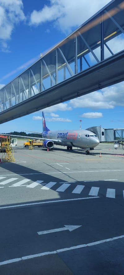 This image, captured from inside a jet bridge, depicts an airport scene at Khrabrovo, Russia, on a bright, partially cloudy day. The primary subject is a passenger aircraft, identifiable as a Smartwings Boeing 737, connected to the jet bridge.

The aircraft features a silver-grey fuselage, with its nose pointing towards the left of the frame (if the image were rotated to portrait). Prominently displayed on its side in orange and red lettering is the airline's name, "smartwings". The tail fin and engine nacelles are a dark blue or purple, consistent with the Smartwings livery. The jet bridge extends from the left of the frame, partially obscuring the aircraft's wing and the terminal building behind it. Its transparent roof and side panels allow views of the bright blue sky with fluffy white clouds overhead.

Below the aircraft, the dark grey tarmac is visible, marked with various white, red, and yellow lines. A large white arrow points forward on the tarmac, and a 'zebra crossing' style marking is also present. Ground support equipment, including a yellow tug vehicle and possibly scaffolding or maintenance platforms, can be seen near the aircraft's fuselage and engines, suggesting ongoing ground operations such as baggage loading or aircraft servicing. In the distance, a small patch of green trees marks the edge of the airfield. The perspective is from inside the jet bridge, looking out, indicating passengers are likely boarding or disembarking the aircraft.