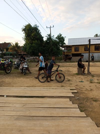 The image captures an outdoor scene in what is identified as the city of Turumame, Indonesia. The foreground features a wooden walkway constructed from rough-hewn planks, with patches of dry grass and dirt visible between them. Beyond the walkway, the ground is predominantly sandy and uneven, with sparse vegetation.

In the middle ground, several individuals are present. A young boy on the left is standing with a BMX-style bicycle, its frame black and red, and its wheels are black. He appears to be holding onto the handlebars. To his right, a young man wearing a white and blue striped t-shirt and black shorts walks away from the camera. Further behind them, two young boys are seated on a scooter, one in front of the other. Several motorcycles are parked to the left of the scene.

In the background, there are a few buildings. The most prominent is a two-story wooden structure with a corrugated metal roof, painted a light yellow color, featuring numerous windows with dark frames. To the left of this structure, another building with a darker roof and visible support beams is partially obscured by trees. Tall trees with dense green foliage are scattered throughout the area, and utility poles with overhead wires are visible against the sky. The sky is partly cloudy with hues of blue and white, suggesting it might be late afternoon or early morning. Two men are seated on a wooden block near the yellow building, and another boy stands further in the background. Several pairs of flip-flops are scattered on the ground. The overall impression is a casual, community-oriented setting.