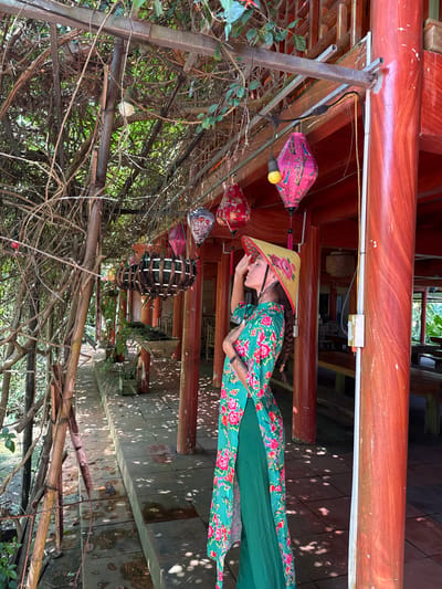 An outdoor scene captures a person in traditional Vietnamese attire within a covered walkway or veranda, likely in Thông Nguyên Commune, Vietnam. The central subject, positioned on the right, is a person wearing a vibrant green áo dài adorned with red and pink floral patterns. They are also wearing a traditional Vietnamese conical hat (nón lá) decorated with matching flowers, and their hair is styled in a braid. The person stands with one hand on their hip and the other raised towards their face/hat, looking slightly upwards.

The setting features prominent red-stained wooden beams and pillars overhead, indicative of traditional architecture. A series of colorful lanterns, including pink ones with bird motifs, red ones with floral designs, and a woven wooden lantern, hang from a metallic and bamboo overhead structure. The tiled ground beneath shows dappled sunlight and distinct shadows, suggesting a bright, sunny day. The left side of the frame is dominated by a dense growth of vines and branches, forming a natural archway or canopy. There is no visible text in the image.