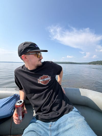 A man wearing a black Patagonia baseball cap, yellow-tinted sunglasses, and a black t-shirt with a Harley-Davidson logo is sitting in an inflatable boat on a body of water. He is holding a red and silver can in his right hand. The boat is moving on a calm lake, with a forest-covered shoreline in the background. The sky is blue with scattered white clouds. The overall scene depicts a leisurely day out on the water. This location is the Krasnoyarskoye reservoir near the city of Divnogorsk, Krasnoyarsk Krai, Russia, not Sylva.