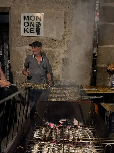 The image depicts an outdoor grilling scene, likely in Santiago de Compostela, Spain, given the context. In the foreground, a man in a grey polo shirt and a black cap is standing by a grill laden with what appear to be small fish or seafood. He is looking off to the side with a slight smile. Multiple metal grill racks are visible, some already holding food and others empty, placed over a charcoal fire that emits a faint haze. Behind the man, a stone wall serves as the backdrop, adorned with a sign that reads "MON ONO KE" with a stylized character. To the right of the wall, a vertical pipe is covered in various stickers. The overall atmosphere suggests a casual outdoor food stall or restaurant setup, possibly during an evening or nighttime event, given the lighting.