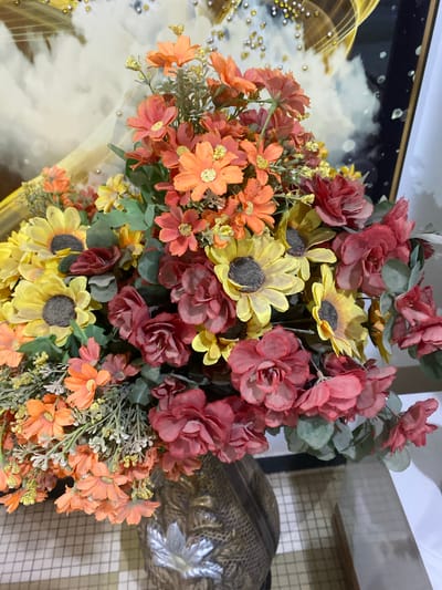 This is a close-up, eye-level, vertical, medium shot of a vibrant artificial flower arrangement in a decorative vase. The bouquet is a mixture of sunflowers, roses, and smaller daisy-like flowers in shades of yellow, orange, and red, interspersed with green foliage. The vase itself appears to be made of a textured, metallic material with an ornate pattern. In the background, a framed piece of art with abstract golden and white swirls is visible, along with a white wall. The flowers are the primary focus, filling most of the frame. There are no people or animals present, and no actions are taking place. The scene is indoors, likely in a home or decorative setting. The information about Giginyu, Nigeria is not supported by the visual content of the image.