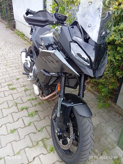 A black BMW F900XR motorcycle is parked on a paved sidewalk in Wilczyce, Poland. The motorcycle is facing forward and is positioned slightly to the right of the frame. It has a tall windscreen, handguards, and a prominent exhaust pipe. The motorcycle is clean and appears to be in good condition. The sidewalk is made of interlocking concrete pavers and has some weeds growing between them. To the right of the motorcycle, there is a dense green bush with small yellow flowers. Behind the motorcycle, a light-colored wall and a fence are visible. The lighting suggests it is daytime. The date stamp "09.06.2026 12:49" is visible in the lower right corner of the image. There are no people or animals in the frame. The overall scene depicts a parked motorcycle in an outdoor urban or suburban environment.