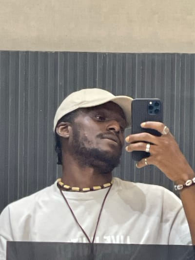 A selfie of a Black man wearing a light-colored baseball cap and a white t-shirt is reflected in a mirror. He has a beard and is wearing a necklace with wooden beads and a bracelet made of seashells. He is holding a black smartphone in his right hand, which is raised to take the photo. The background consists of a ribbed, dark gray wall and a lighter-colored wall above it. The image is taken indoors, likely in a bathroom or changing room. It is not possible to determine the exact location in Guzape, Nigeria from the image alone.