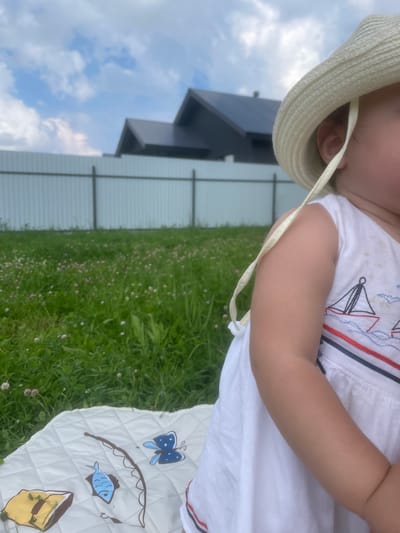 A close-up shot shows a child from the chest down, wearing a white dress with embroidered sailboats and a straw hat, sitting on a quilted blanket. The blanket has embroidered images of a fishing rod, a fish, a butterfly, and a yellow object. The child is outdoors in a grassy area with small pink and white flowers. In the background, a white metal fence separates the lawn from two dark-roofed houses. The sky above is blue with white clouds.
