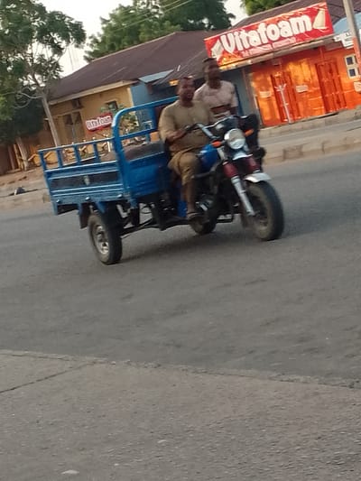 In Pantame, Nigeria, two individuals are riding a blue three-wheeled motorcycle. The motorcycle has a cargo bed at the rear. The driver, wearing a light brown shirt and khaki pants, is operating the vehicle. A passenger, wearing a patterned shirt, is seated behind the driver. They are on a paved road, with buildings and trees visible in the background. A red and white sign with the word "Vitafoam" is prominently displayed on one of the buildings. The scene appears to be during daylight hours, possibly late afternoon due to the lighting.