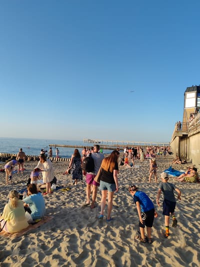 A crowded beach scene unfolds under a clear blue sky, likely during the late afternoon given the warm, low-angled sunlight casting long shadows across the sand. The ocean, a calm expanse of blue, stretches to the horizon where a pier with a long walkway is visible, backed by a line of wooden posts in the water.

Numerous people are scattered across the sandy beach, engaged in various beach activities. Some are in the shallow water, swimming or wading, while others sit on towels or lie on the sand. Groups of people are standing and conversing, and individuals are seen walking along the shoreline. Children are also present, some playing in the sand or near the water's edge.

Notable details include the presence of beach towels, bags, and a blue inflatable toy. The architecture of a building with a prominent glass facade is visible on the right side of the image, suggesting a developed beachfront area. The overall atmosphere appears relaxed and summery, with people enjoying a pleasant day at the seaside. The location is identified as Zelenogradsk, Russia. There is no visible text in the image.