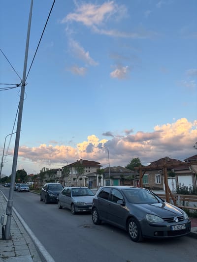 This image depicts a residential street scene in the city of Popovo, Bulgaria, captured during late afternoon or early evening. The perspective is from a low angle, looking down the street, with a significant portion of the frame dedicated to the sky.

In the foreground, on the left, a tall, light-gray utility pole stands vertically, adorned with multiple black electrical cables and connection boxes. A paved sidewalk, showing some wear and interspersed weeds, runs parallel to the pole. Adjacent to the sidewalk is an asphalt road, marked by a solid white line defining the parking area.

Several cars are parked along the right side of the road, facing generally toward the left of the frame. The most prominent vehicle in the foreground is a dark gray Volkswagen Golf (fifth generation) hatchback, positioned close to the curb. Its front license plate, "B 3252 TP," indicates a Sofia registration. Behind it, a silver Peugeot 306 hatchback is parked. Further down the street, a dark gray SEAT Ibiza hatchback is visible, followed by a silver car partially obscured by the utility pole, and a white car in the distance.

On the right side of the road, behind the parked cars, a series of two-story residential buildings line the street. These structures feature tiled roofs, some with visible chimneys. Architectural details include windows with varying frames, some appearing green or light blue. One building has a prominent wooden pergola or decorative structure attached to its facade. The overall architectural style suggests typical Eastern European residential construction.

Green foliage from trees and bushes is visible along the street, interspersed between the buildings and providing natural elements to the urban environment.

The upper two-thirds of the image are dominated by a clear blue sky, transitioning to warmer, pinkish-orange hues near the horizon. Distinct, fluffy cumulus clouds are present, illuminated by the setting sun, particularly pronounced above the buildings on the right. Smaller, more dispersed clouds are scattered across the higher parts of the sky. The lighting is soft and diffused, characteristic of twilight hours. No individuals are visible in the scene.