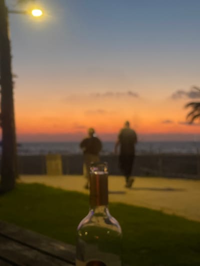 The image depicts a blurred, low-light scene consistent with a sunset over the Mediterranean Sea from Herzliya, Israel. In the immediate foreground, the top of a glass bottle, likely containing a light-colored liquid, is centrally positioned on a dark, horizontally slatted surface, presumably a wooden table or railing. To the left, a patch of dark green grass is visible. Beyond the foreground, two indistinct, dark figures are seen walking away from the camera on a paved promenade or path. To their left, a dark vertical structure, possibly a tree trunk or lamppost, is partially visible. Further right, blurred dark foliage, characteristic of palm trees, can be observed. The midground features a dark, horizontal barrier separating the path from the body of water. The sea itself is visible, showing subtle texture. The background is dominated by a vibrant sunset sky, transitioning from a light blue at the top to intense orange and red hues near the horizon, with several indistinct cloud formations. A single, bright yellow-white light source, indicative of a streetlamp, is present in the upper left portion of the frame. The overall impression is an evening view from a beachfront establishment, overlooking the promenade and sea.