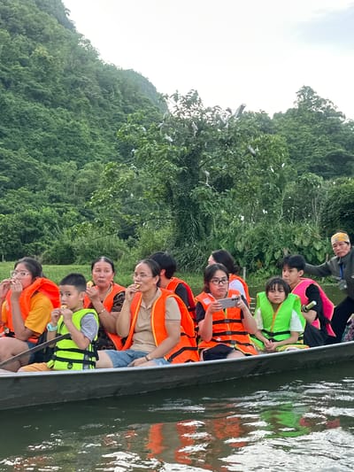 In the city of Yên Sơn Ward, Vietnam, a group of people are on a boat tour. Several individuals, including children and adults, are wearing orange and green life vests. The boat is filled with passengers who appear to be enjoying a leisurely outing. In the background, lush green trees cover a mountainside, with a flock of birds perched on branches. The water in the foreground reflects the boat and its passengers, creating a serene and picturesque scene.