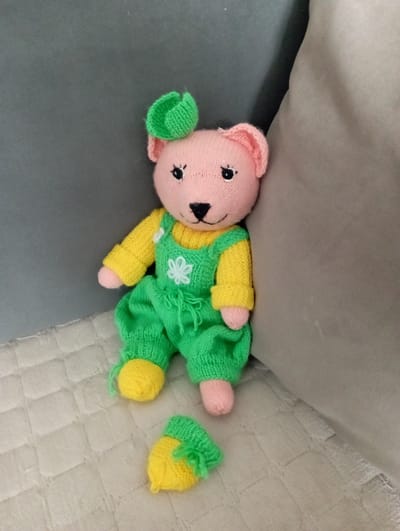 The image shows a pink knitted teddy bear lying on its side on a textured, light-colored surface, possibly a couch or bed. The bear is wearing a yellow long-sleeved shirt and green overalls. The overalls have yellow trim at the cuffs and legs, and a white knitted flower embellishment on the chest. The bear's ears, nose, and paws are also knitted in a pink or nude color. Two small, knitted yellow items with green trim, resembling hats or booties, are placed to the right of the bear. The lighting is soft, suggesting an indoor setting during the daytime. The overall scene is calm and domestic, with no text visible. The location context is Ulan-Ude, Russia.