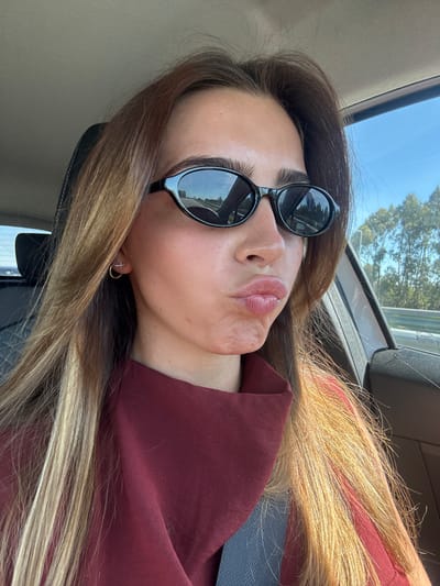 A female subject is depicted in a medium close-up selfie inside a vehicle, likely on a road within the Aveiro, Portugal region. She has long, multi-toned brown hair with prominent blonde highlights. Her face is directed towards the camera, with lips pursed in a "kiss" pose. She wears black, oval-shaped sunglasses that obscure her eyes. Two small, gold-colored earrings are visible on her right ear: a small hoop and a cross-shaped stud. Her attire consists of a dark red garment, possibly a top, with visible folds around the neck area. A portion of a blue fabric is visible below the red garment on the lower left. The vehicle's interior features a light gray headliner and a black headrest. Outside the car, visible through the right window and reflected in her sunglasses, is a clear blue sky, a road with a guardrail, and trees with light-colored trunks, consistent with a sunny daytime environment.