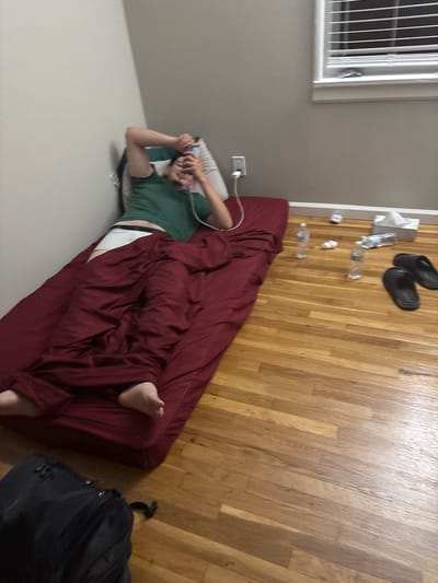 An indoor scene captured from a high, tilted angle shows a room with light gray walls and light-colored hardwood flooring. A person, identified as male, is lying on a maroon mattress or futon placed directly on the floor. He wears a green t-shirt and white underwear/shorts, with a maroon sheet or blanket partially covering his lower body. His head rests on a light-colored pillow, and he is holding a smartphone, looking at it with a slight smile, suggesting a relaxed or amused state. A white power outlet with a plugged-in white cable is visible on the wall near his head.
The floor next to the mattress is somewhat cluttered with everyday items: a pair of black sandals/slides, several empty or partially empty plastic water bottles, crumpled tissues or paper, and a white paper bag. In the upper left corner of the frame, a window with white horizontal blinds is visible. A black backpack rests on the hardwood floor in the lower right corner of the image. The setting appears to be a casual living space, possibly a bedroom, in Franklin Township, United States of America. No specific time of day or weather conditions are discernible from the indoor lighting. There is no visible text in the image.