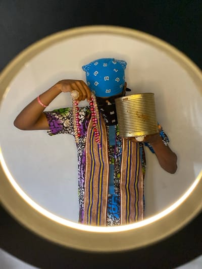 A partially visible individual stands within the circular illumination of a ring light against a plain white background, indicative of an indoor setting in Ngleshi Amanfro, Ghana. The person wears a blue bandana with white patterns covering their head, a black face mask obscuring their lower face, and a multi-colored shirt featuring purple, green, blue, and white batik-style patterns, with two vertical strips of woven fabric in brown, blue, and yellow stripes. A pink wristband is visible on the left wrist. The left hand displays a prominent gold-colored, mesh-patterned ring on the ring finger and holds multiple strands of pink and clear beaded necklaces. The right hand supports a cylindrical, ribbed metal can with a slightly tarnished, golden hue. The person's face is largely obscured.