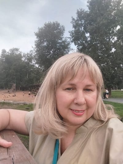 The image is a selfie taken outdoors at a sandy beach or park area with scattered trees and a cloudy sky. In the foreground, a blonde woman with fair skin and rosy cheeks smiles at the camera, wearing a light tan collared shirt. Her left arm is resting on a wooden railing. In the background, there are several people scattered on the sand, some appearing to be sunbathing or interacting. There is also a wooden structure with a playground-like element visible. The overall setting suggests a recreational area within a city. While the specific location within Orenburg, Russia, is not visually identifiable, the scene depicts a typical outdoor leisure space that could be found in many cities.