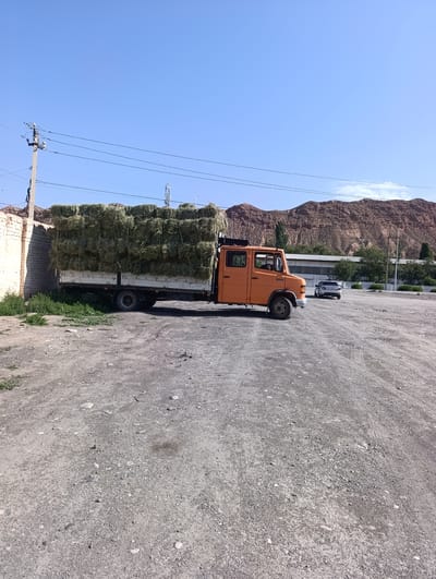 An orange truck is parked on a gravel lot. The truck is loaded with large bales of hay. The truck is facing to the right. A white car is parked behind the truck, also facing to the right. In the background, there are mountains, trees, and buildings. The sky is clear and blue. The ground is covered in gravel and dirt. The scene appears to be in a rural area. The image is taken from a slightly elevated angle.