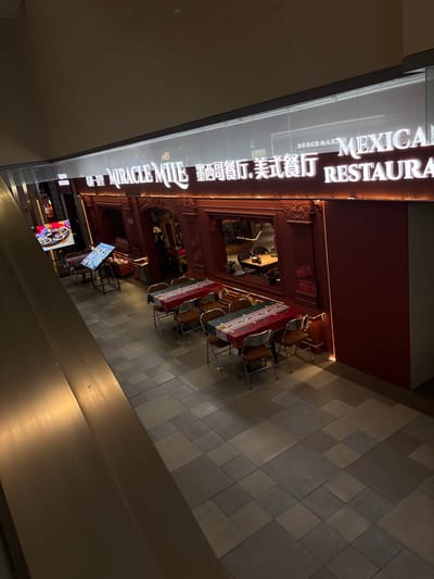 An overhead, high-angle shot captures the exterior of a Mexican restaurant named "MIRACLE MILE MEXICAN RESTAURANT" in Heping District, China. The restaurant's facade is painted a vibrant red, with decorative moldings around windows and entrances. A large, illuminated sign above the entrance displays the restaurant's name in both English and Chinese characters. Inside, tables are set with festive, patterned tablecloths, suggesting a holiday theme, possibly Christmas, due to the red and green colors and snowflake patterns. Chairs with metal frames surround the tables. To the left, a digital display screen showcases food items. The restaurant floor is tiled in a geometric pattern of beige and gray squares. The surrounding environment appears to be an indoor shopping mall or a similar commercial space, with a metallic railing visible in the foreground. The lighting is artificial, creating a warm and inviting ambiance within the restaurant. There are no people visible in the immediate scene.