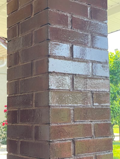 The image shows a close-up of a dark brown painted brick pillar. The paint appears to be glossy and has a wet or streaky appearance in some areas, suggesting it may have been recently applied or exposed to moisture. The bricks are laid in a standard running bond pattern. The pillar is positioned outdoors, as indicated by the presence of green foliage and what appears to be a rose bush with red flowers in the background to the left. A portion of a white soffit and a downspout are visible on the left side of the frame, suggesting the pillar is part of a building's exterior structure, possibly a porch or entryway. The lighting suggests it is daytime. The location is stated as Slobozia-Dușca, Moldova. No people are visible in the image.