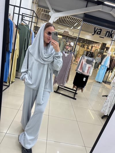 A person is taking a selfie in a clothing store. They are wearing a light blue tracksuit and a matching headscarf, with sunglasses. The store appears to be well-lit and organized, with racks of clothing visible in the background. The floor is tiled, and the overall impression is one of a modern retail environment. There are no obvious indications of the time of day or weather outside. The person's expression is neutral. Visible text includes "Ya nk" on a sign in the background. The location is identified as Nizhnekamsk, Russia.