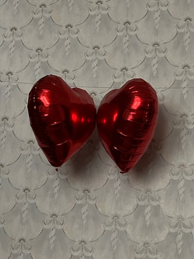 Two red heart-shaped balloons are suspended against a white, textured wall with a repeating floral pattern. The balloons appear to be made of foil and are slightly deflated, with visible creases. They are positioned close together, touching or nearly touching in the center. The lighting creates reflections on the shiny surface of the balloons. The background wall has a raised, embossed design. There are no people or specific landmarks visible in the image. The scene suggests a celebratory or decorative context, possibly for a holiday like Valentine's Day or a birthday. No information about the city of Synelnykove, Ukraine, can be inferred from this image.