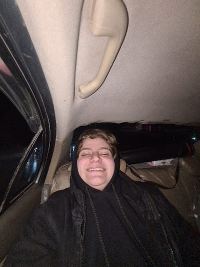 An image captured from inside a car shows a smiling individual. The person is wearing a black hooded jacket. A beige car interior with a handle is visible to the left. The car window is partially visible at the bottom, offering a glimpse of a dark exterior. A few boxes can also be seen behind the smiling person.