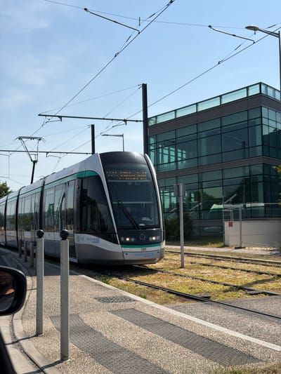 The image shows a tram stopped at a station in Rungis, France. The tram is modern and sleek, with a white and gray exterior and large windows. It is positioned on tracks that run alongside a grassy area and a concrete platform. In the background, a contemporary glass-fronted building stands tall under a clear blue sky, suggesting a bright and sunny day. Overhead, electrical wires for the tram system crisscross the frame.
The tram's destination display shows "707" and "ile de France mobilités," indicating it's part of the public transportation network in the Île-de-France region. The time of day appears to be daylight, likely late morning or early afternoon, given the bright lighting and clear sky. The overall scene is urban and functional, depicting a common mode of transportation in a modern city setting. No specific activity or event is explicitly shown, other than the tram being at a stop, implying passengers may be boarding or alighting. The image is oriented vertically, suggesting it was taken from a perspective looking up at the tram.