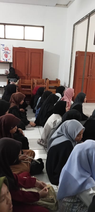 The image shows a group of women in black hijabs sitting on the floor in a room in Cangkuang, Indonesia (latitude: -7.065, longitude: 107.552). There are four wooden chairs, two bookshelves with double doors, and a poster on the wall. The women are engaged in a discussion. One woman in the center of the frame is wearing a light blue hijab and is looking down at her hands, while the woman to her right is wearing a pink hijab. There is a white tile floor, and there are numerous bags and belongings scattered around. There is a window with two panes. The room has a white ceiling with light fixtures.