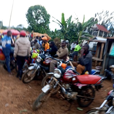 The image depicts a group of people gathered around several motorcycles in what appears to be an outdoor area. It seems to be at Borabu Chitago ward, Kenya. There are about 10 people, some standing and some sitting on the motorcycles. One man is wearing a blue jacket, one is in a beige jacket, one is wearing an orange cap, and another is in a black jacket. The motorcycles are a mix of red and white, and several people are sitting on them. The ground appears to be dirt or mud. There is a tree and banana tree in the background, as well as what appears to be a corrugated metal structure.