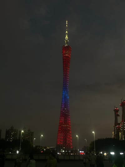 The Guangzhou Tower, also known as the Canton Tower, is lit up in red and blue lights, in the city of Guangzhou Shi, China, at night.  The coordinates for the tower are latitude: 23.117, longitude: 113.319. The tower appears to be a popular tourist destination, as numerous people and streetlights are visible in the foreground.  There are some building structures obscured by foliage in the bottom right corner of the image, which are likely residential. The sky is mostly dark, with a few clouds visible in the distance.  The tower is the main focal point of the image, with its intricate lattice structure and illuminated colors making it the most prominent feature of the scene.
