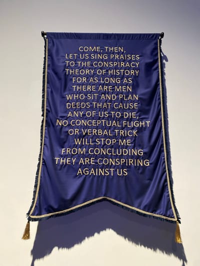 The image shows a dark blue banner with gold embroidered text, hanging from a rod against a white wall. The text reads: "COME, THEN, LET US SING PRAISES TO THE CONSPIRACY THEORY OF HISTORY FOR AS LONG AS THERE ARE MEN WHO SIT AND PLAN DEEDS THAT CAUSE ANY OF US TO DIE, NO CONCEPTUAL FLIGHT OR VERBAL TRICK WILL STOP ME FROM CONCLUDING THEY ARE CONSPIRING AGAINST US." The banner has gold fringe along its bottom edge and gold trim along its sides. Two gold tassels hang from the bottom corners of the banner. The lighting casts a shadow of the banner and its tassels on the wall. The image is an uncropped, medium shot taken from a slightly low angle. The location is not specified.
