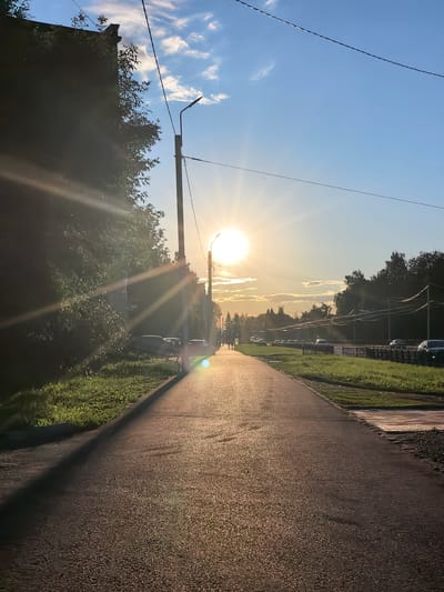 The image captures a sunlit pedestrian path in Sterlitamak, Russia, during what appears to be late afternoon or early evening. The sun, low in the sky, casts a warm glow and creates lens flares that streak across the scene. A paved pathway runs through the center of the image, leading the eye towards the horizon. To the left of the path is a dense line of trees and the side of a building, partially obscured by foliage. To the right, a road with parked cars and oncoming traffic is visible, separated from the pedestrian path by a grassy strip and a fence. In the distance, more trees form a dark silhouette against the bright sky. The sky itself is a clear blue, with a few wispy clouds scattered near the sun. A street lamp stands near the building on the left, and power lines stretch across the upper portion of the image. Two small figures can be seen walking in the distance on the pedestrian path.