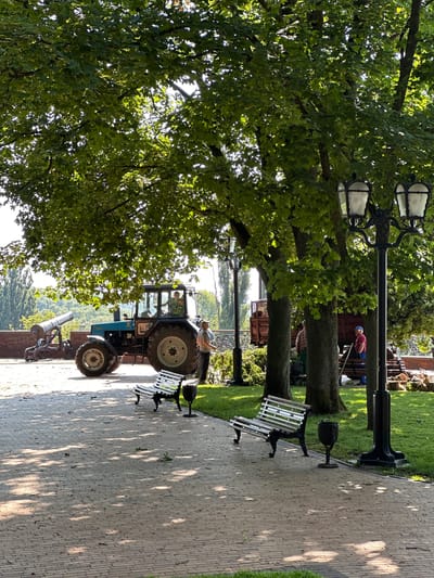 The image depicts an outdoor scene in Chernihiv, Ukraine, likely a park or historical site. In the foreground, a paved pathway with a brick pattern is visible, with two empty park benches casting shadows from the overhead trees. The pathway is bordered by a manicured lawn.
Dominating the mid-ground is a blue tractor, positioned on the paved area. To the left of the tractor, a vintage cannon on a wooden carriage is visible, resting on a red brick wall that delineates a slightly elevated area. Several people are present around the tractor and in the background, seemingly engaged in some activity. One individual in a camouflage uniform is seated in the tractor's cabin, while another person in a grey shirt and cap stands near the rear of the tractor. Further back, two individuals in casual clothing are seen near a wooden trailer.
The background features mature trees with lush green foliage, creating a canopy that filters the sunlight and casts dappled shadows. In the distance, more trees and a glimpse of buildings or structures can be observed. The overall atmosphere suggests a warm, sunny day. The presence of the cannon and the tractor may indicate a historical reenactment, a local event, or a display of historical machinery.