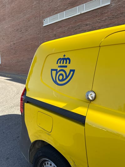The image shows a close-up of the side of a bright yellow vehicle, possibly a van or bus, parked outdoors. The vehicle has a dark gray trim along its side and a black rubber element at the bottom, partially obscuring a tire. A circular metallic fuel cap with a lock mechanism is visible on the yellow panel. A blue logo, which appears to be a stylized "P" combined with some circular elements, is prominently displayed on the side of the vehicle. To the left of the vehicle, a section of a brick wall with a white slatted grate is visible. The scene suggests it is daytime, with sunlight casting shadows, and the ground appears to be asphalt. The overall impression is of a vehicle parked in an urban or industrial setting. The image appears to be taken in Calella, Spain, although there are no explicit text labels or signs to confirm this within the frame. No people are visible in the image, and no specific activity or event is depicted. The focus is solely on the vehicle and its immediate surroundings.
