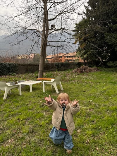 The image displays an outdoor garden scene in Erba, Italy, under an overcast sky.
A young child, approximately a toddler, is centered in the lower foreground, kneeling or squatting on green grass. The child has blonde hair and a dark pacifier in its mouth. It is dressed in a beige corduroy puffer jacket, a dark green long-sleeved shirt, blue pants patterned with the text "Bobo Choses," and light brown shoes. Both arms are raised with open hands, fingers splayed.
In the midground, a prominent deciduous tree with bare branches stands centrally. A gray and white tabby cat is perched on one of its upper branches. To the left of the tree, a white rectangular plastic table is accompanied by two matching white plastic chairs. Two gray plant pots are situated near the left chair. An orange metal bench with a woven pattern is visible behind the table. To the right of the central tree, a large dark green coniferous tree and a smaller palm tree are present. A low wall covered in ivy runs across the background behind the garden furniture.
Beyond the wall, residential buildings with orange-tiled roofs are visible. The distant background features a range of hazy mountains under the overcast sky. A construction crane is faintly discernible on the left side of the mountain range. A pile of brown leaves and debris is located on the grass in the right midground.