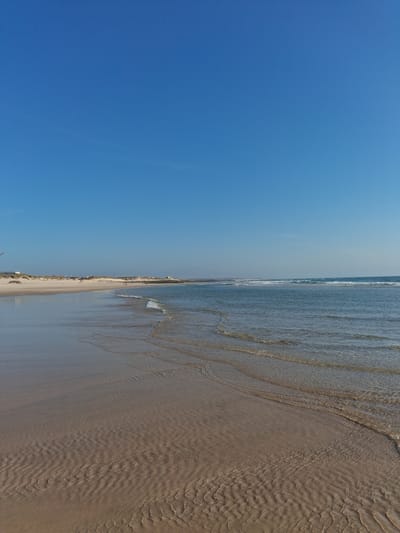 The image depicts a beach scene, likely taken during the day. The right side of the image showcases a sandy beach with water gently lapping at the shore, creating rippled patterns in the sand. On the left, a clear blue sky dominates, suggesting good weather conditions.
Based on the mention of Figueira da Foz, Portugal, this is likely the Praia da Claridade. This beach is known for its long stretch of sand and its urban setting. The lack of specific landmarks or people makes it difficult to provide a more detailed description of the location or any particular activities taking place.