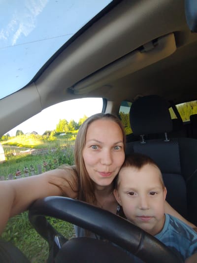 A woman and a young boy are inside a car, with the woman in the driver's seat and the boy in the passenger seat. The woman is smiling and looking at the camera, while the boy is looking forward. The car's interior, including the dashboard and seats, is visible. Outside the car, a rural landscape with green trees and houses can be seen, suggesting a daytime setting. The sky is bright, indicating fair weather. The scene appears to be in Чухлэм, Russia.
