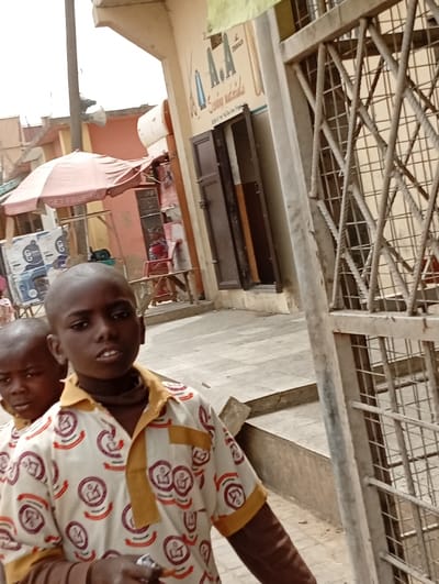 In Gawuna, Nigeria, two young males are captured outdoors in what appears to be a commercial street. The foreground shows a male with a shaved head and dark complexion, wearing a white short-sleeved shirt patterned with reddish-brown circular motifs and featuring mustard yellow collar and sleeve cuffs. A dark fabric is visible around his neck, and he holds a small, light-colored object in his left hand. Partially obscured behind and to the left of him is a second male, also with a shaved head. They stand on a grey paved surface, with concrete steps visible behind them. To the right, a weathered metal gate with a diamond lattice pattern partially frames the scene. In the background, a light-colored building displays a sign with "A.A Sewing materials" and illustrations of scissors and thread. Its dark brown double doors are partially open. Further to the left, a red market umbrella with "GET FREE" text and various items are visible, suggesting a street vendor's stall.