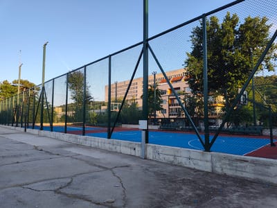 The image shows an outdoor basketball court in Svishtov, Bulgaria. The court has a blue playing surface with white markings and red outer sections. It is enclosed by a green metal fence. In the background, there is a multi-story building and several trees. The sky is clear and blue. The foreground shows a cracked concrete surface. There are no people visible in the image. The overall scene depicts an empty sports facility.