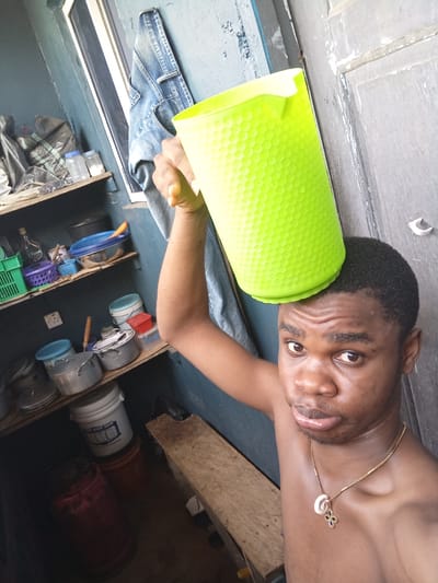 A shirtless young man stands in what appears to be a domestic indoor space, possibly a kitchen or storage area, identified through visible items such as pots, buckets, and shelving along a wall. The man holds a bright green, textured plastic cup/container on his head with his right arm raised. He is looking directly at the camera. He is wearing a gold chain with a pendant around his neck. He is in Owerri, Nigeria. The room is small and cluttered. Light is coming from the window.