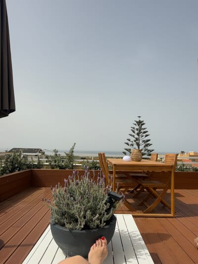 The scene captures a rooftop terrace overlooking a cityscape in Vila Nova de Gaia, Portugal, under a hazy, overcast sky. The terrace is furnished with wooden decking, a light-colored wooden table, and chairs. A potted plant with lavender sits on a white bench. A Norfolk pine stands on the table. A woman's foot with red nail polish is visible, suggesting someone is relaxing on the bench. The city's buildings and rooftops stretch into the distance towards the sea.