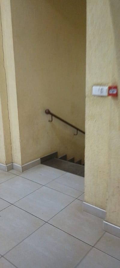 The image depicts an indoor stairwell area in a building located in Ngong ward, Kenya. The scene is illuminated, with no visible light source. The walls are painted a light beige or yellow, exhibiting a slightly textured finish and some vertical cracking on the left structural pillar. The floor consists of light grey square tiles with dark grout lines. A dark grey or brown staircase descends from the foreground towards the lower right, partially obscured by a wall section on the right. A dark brown or metallic handrail, supported by two visible brackets, runs along the left wall adjacent to the descending stairs. Light grey skirting boards are installed at the base of all visible walls. On the right wall, two rectangular wall-mounted devices are present: a white unit with indiscernible markings to the left, and a red unit to its right, consistent with an emergency break-glass or pull station. No individuals or active subjects are present in the frame.