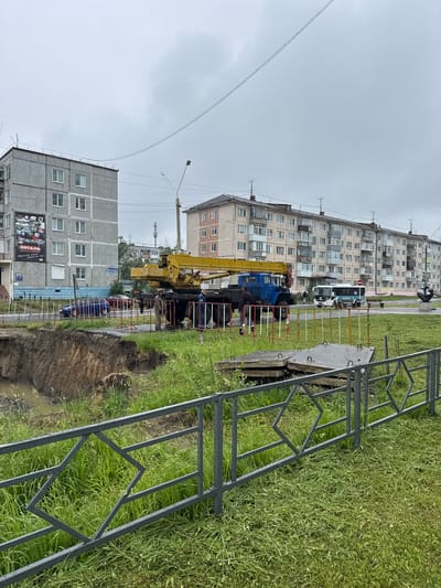 The image depicts a scene in Usinsk, Russia, focusing on the aftermath of a land collapse. In the foreground, a metal fence with a diamond-shaped pattern separates a grassy area from a significant land erosion site. The collapsed area reveals a steep, muddy bank with standing water at its base.

In the mid-ground, a yellow crane truck and a blue truck are positioned near the edge of the collapse. Two construction workers are visible near the trucks; one is wearing a brown uniform, and the other is in a blue uniform. A white bus with the word "УСИНСК" (Usinsk) visible on its side is driving away from the collapse site.

The background features several multi-story apartment buildings characteristic of Soviet-era architecture. One building on the left has a large advertisement poster on its facade. Power lines crisscross the sky, and the overall atmosphere suggests an overcast or rainy day. The environment appears to be a residential area undergoing some form of construction or repair work related to the land collapse.