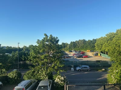 In Schweitenkirchen, Germany, on a clear day with a vibrant blue sky, a parking lot is visible. The lot contains a variety of cars, including silver sedans, a red hatchback, and several other vehicles in various colors and models. The parking lot is bordered by a green fence and lush trees, some of which have white flowers. A street runs alongside the parking lot, with more trees and foliage lining the road. In the foreground, two silver cars are parked. The scene appears to be a typical urban or suburban setting, with a focus on the parked vehicles and the surrounding greenery. There are no people visible in the image.