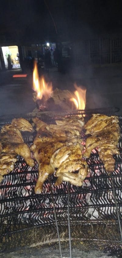 The image captures a nighttime scene of chicken being grilled over an open fire. In the foreground, several pieces of seasoned chicken are laid out on a metal grill grate. The chicken appears to be spatchcocked or butterflied, with visible skin and some charring or browning, indicating it's in the process of cooking. Below the grill grate, glowing red embers are visible, providing heat.
Behind the chicken, towards the upper middle of the frame, significant flames erupt from the fire, casting a warm, orange glow on the surroundings. These flames suggest that either fat is dripping onto the coals or additional fuel is being burned.
The background is dark and out of focus, indicating that the photo was taken at night. Through the blur, various elements can be discerned:
* To the left, there's a brightly lit opening, possibly a doorway or a stall entrance, with some indistinct shapes inside, perhaps merchandise or furniture.
* Further into the background, scattered artificial lights are visible, resembling streetlights or lights from distant buildings.
* Several blurry figures, presumably people, can be seen in the distance, suggesting a public or semi-public outdoor setting like a market, street, or gathering.
* A faint, thin, diagonal line, possibly a wire or string, is barely visible across the top left portion of the image.
* To the left of the main flames in the background, there's a distinct bright orange/red rectangular shape, which could be another light source or a reflective object.
The overall scene depicts an outdoor cooking event, possibly street food preparation or a backyard barbecue, taking place after dark, with the focus on the cooking chicken and the vibrant flames. No discernible text is visible in the image.
