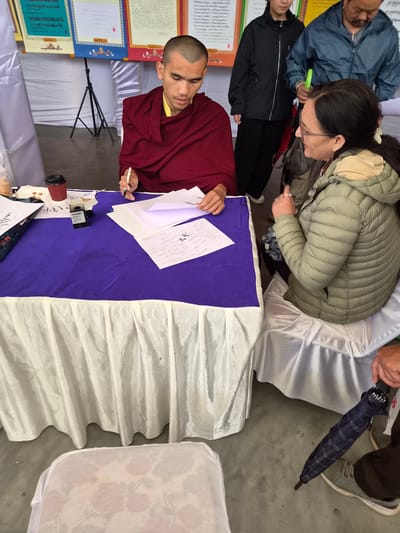 In Dharamshala, India, at an outdoor event, a young Buddhist monk in maroon robes is seated at a table covered with a purple cloth, writing on a piece of paper with a pen. He is focused on his task, with a red paper cup and an ink bottle on the table. A woman wearing glasses and a padded green jacket is seated to his right, looking at the monk. Behind them, a man in a blue jacket stands next to a younger person in a black jacket. Several colorful Tibetan artworks are displayed on a stand in the background. A folded dark blue plaid umbrella is held by a person whose legs are visible in the foreground, indicating a possible break in the weather. The overall scene suggests a cultural or religious gathering.