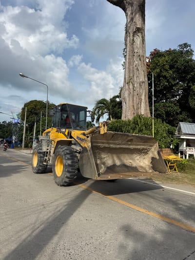 A yellow Komatsu wheel loader is parked on an asphalt road in Ban Tai Subdistrict, Thailand. The loader is positioned on the right side of the road, facing forward. Its bucket is partially filled with dirt and is raised high. The machine has large, thick tires with yellow rims. The driver's cabin has tinted windows, and a person wearing a red shirt is visible inside.

In the background, a very large, old tree with a rough, textured bark dominates the scene. Its trunk is wide and appears to be covered in some vines. To the right of the tree, there is a small building with a metal roof and white walls, with a yellow table and chairs in front of it. Lush green vegetation, including palm trees and other leafy plants, surrounds the tree and the building.

On the left side of the road, a lamppost stands tall. Further down the road, a colorful, tower-like structure with lights is visible, along with some tents or tarps. A person is riding a motorcycle on the road. The sky is partly cloudy with scattered white clouds against a blue backdrop. The road has a yellow dividing line. The overall impression is of an outdoor scene, possibly a roadside, with construction or maintenance equipment present near a natural landmark and some local structures.