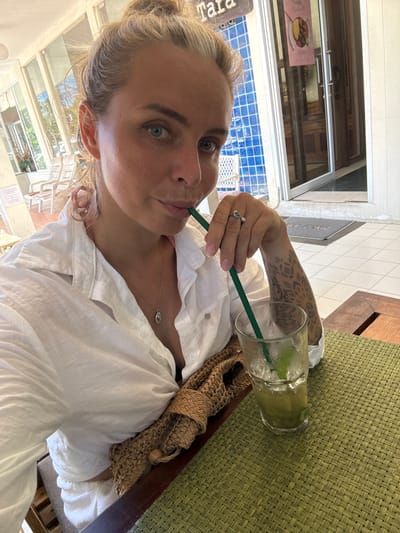A woman with blonde hair pulled back in a bun, wearing a white button-down shirt and a silver necklace, sits at a table outdoors. She is holding a green straw to her lips, about to take a sip from a clear glass filled with a light green beverage, ice, and lime slices. Her right arm, adorned with dark geometric tattoos, rests on the table. She wears a silver ring on her index finger. Beside her, a woven brown bag is placed on the table. The table is covered with a textured green placemat, and a dark wooden chair is visible. In the background, a window with a framed menu can be seen. The scene appears to be in a casual outdoor setting, possibly a cafe or restaurant, with tiled floors and natural light, suggesting it is daytime. The woman's expression is relaxed. Text on a pink menu in the background reads "Nordic Meatballs". The location context is Baan Chaweng Noi, Thailand.